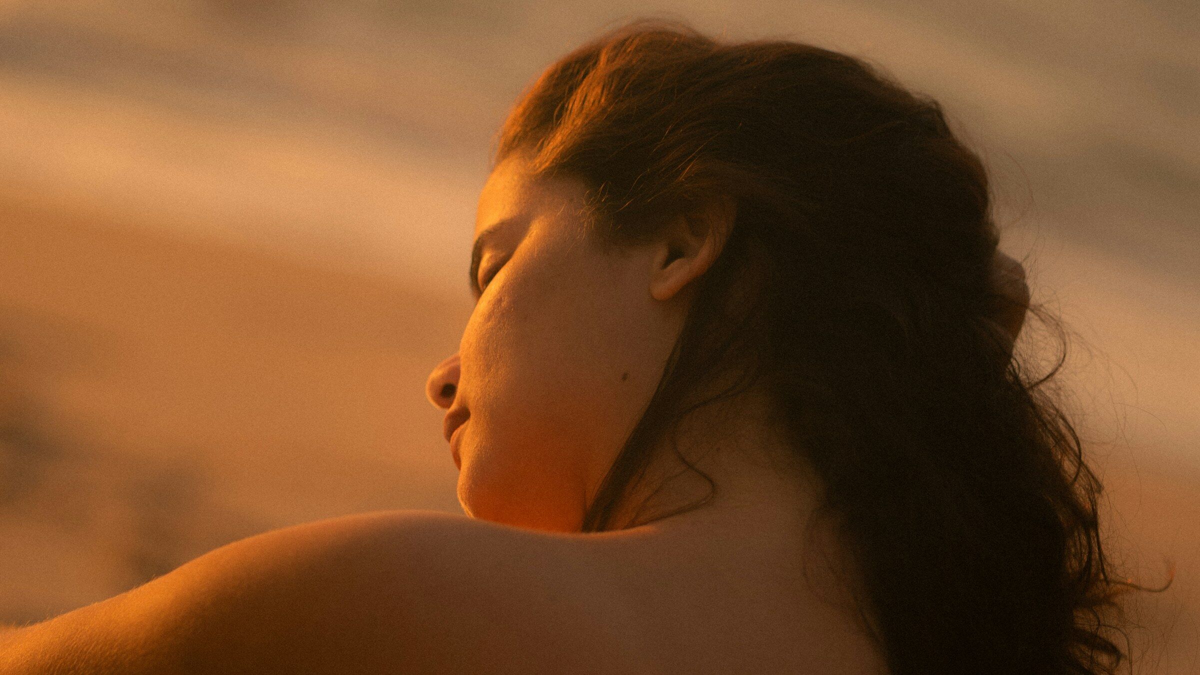 A woman in warm golden light, eyes closed, head turned over her bare shoulder